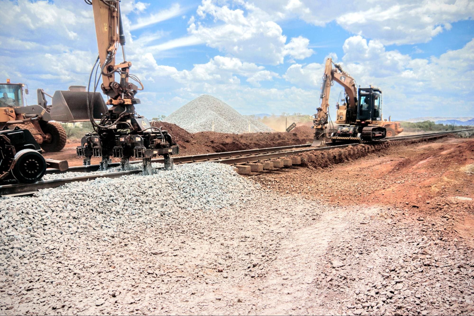 Ballast tamper and ballast undercutter are maintaining rail tracks in Australia near Rio Tinto mines. Red dirt and ballast rock on the ground. Blue skies with bright clouds. 